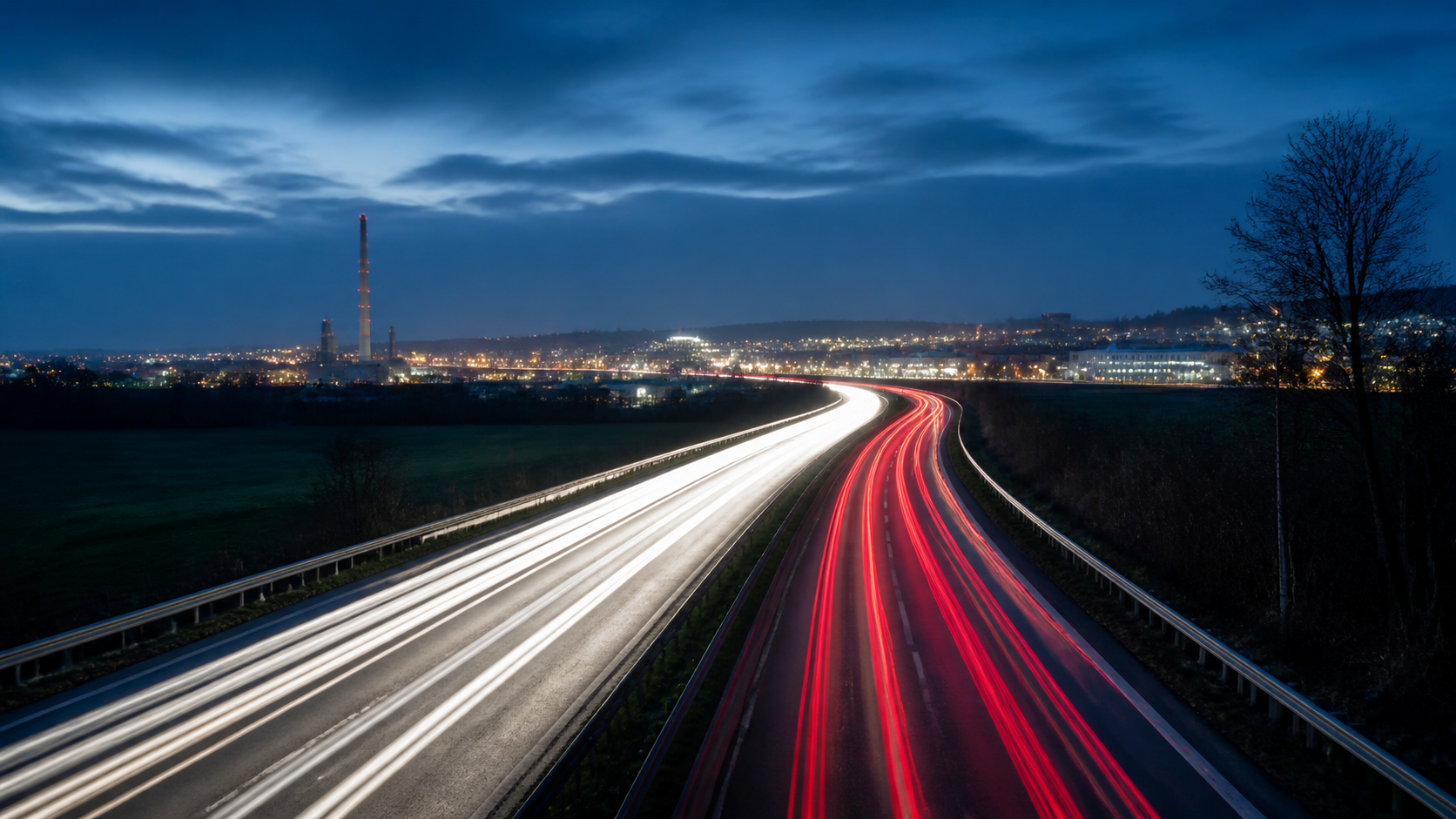 Abendliche Fernstraße mit Lichtspuren in Richtung eines Industriegebiets